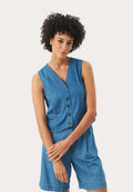 Frau mit lockigem Haar, die eine ärmellose blaue Jeansweste mit Knöpfen und passende Jeansshorts trägt, steht mit verschränkten Armen.