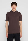Junger Mann steht, trägt ein dunkelbraunes kurzärmeliges Poloshirt und schwarze Hosen vor einem einfarbigen hellen Hintergrund.