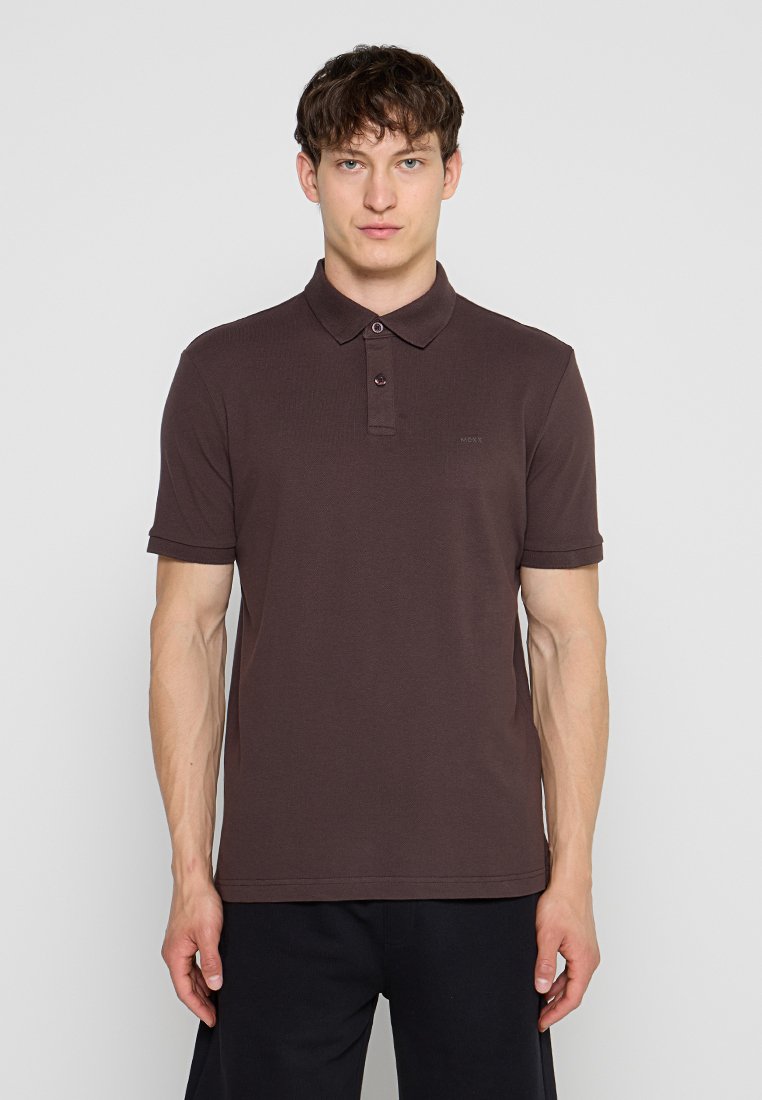 Junger Mann steht, trägt ein dunkelbraunes kurzärmeliges Poloshirt und schwarze Hosen vor einem einfarbigen hellen Hintergrund.