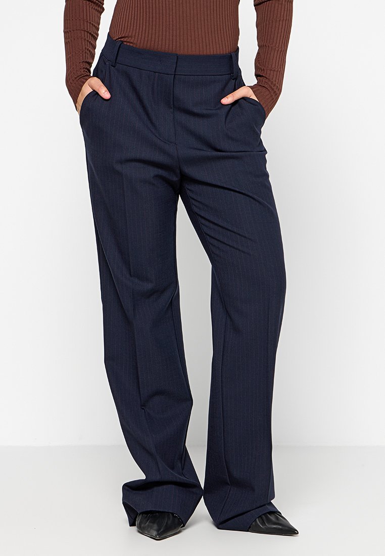 Marineblau gestreifte Hose mit geradem Schnitt, aus einem glatten Stoff gefertigt. Verfügt über Seitentaschen und einen Taillenbund. Getragen mit spitzen schwarzen Schuhen.