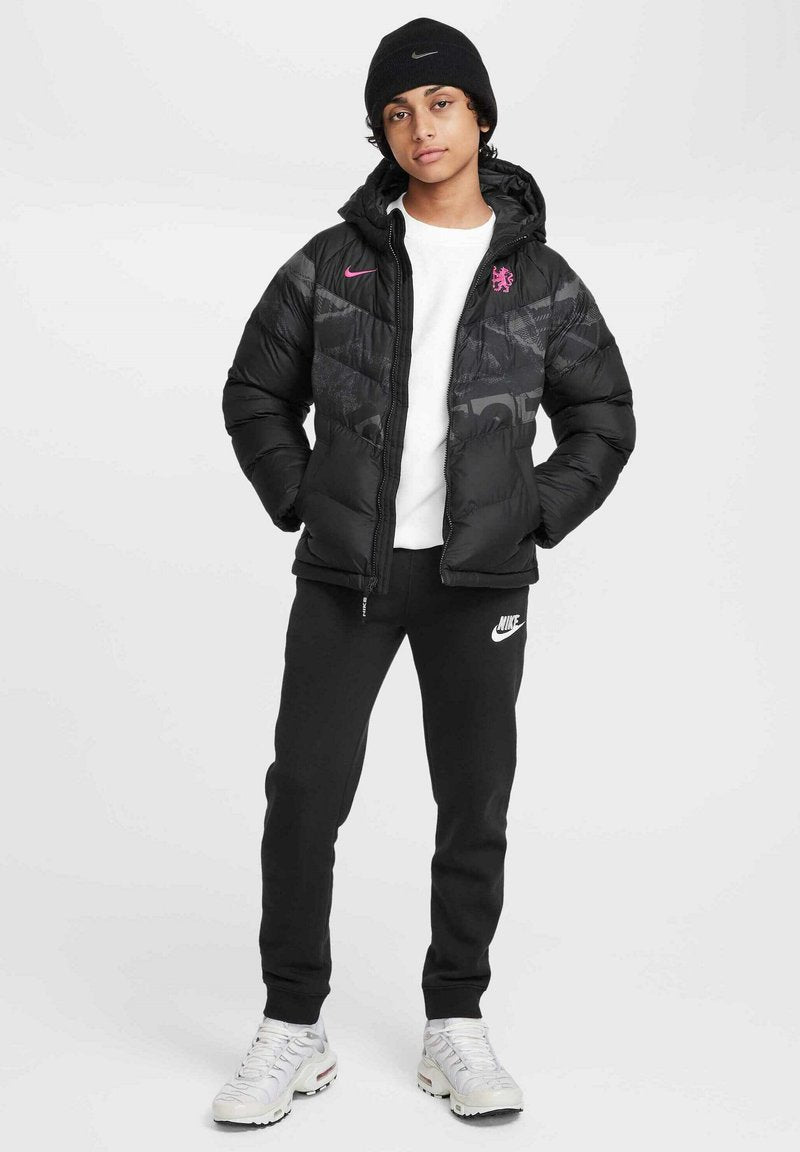 Schwarze Daunenjacke mit gemusterten Akzenten, pinkes Nike-Logo auf der Brust, kombiniert mit schwarzen Sweatpants und weißen Sneakers. Schwarze Mütze auf dem Kopf.