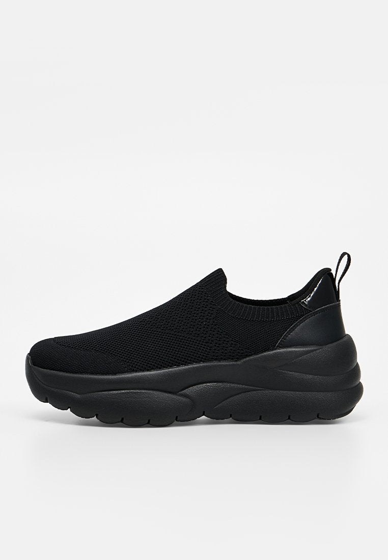 Schwarzer Slip-On-Sneaker mit gestricktem Obermaterial, dicker gepolsterter Sohle und Zuglasche an der Ferse, seitlich auf weißem Hintergrund gezeigt.