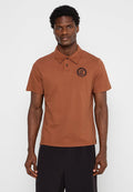 Braunes Poloshirt aus Baumwolle mit Kragen, kurzen Ärmeln und einem runden Logo auf der linken Brust. Glatte Textur und normale Passform.