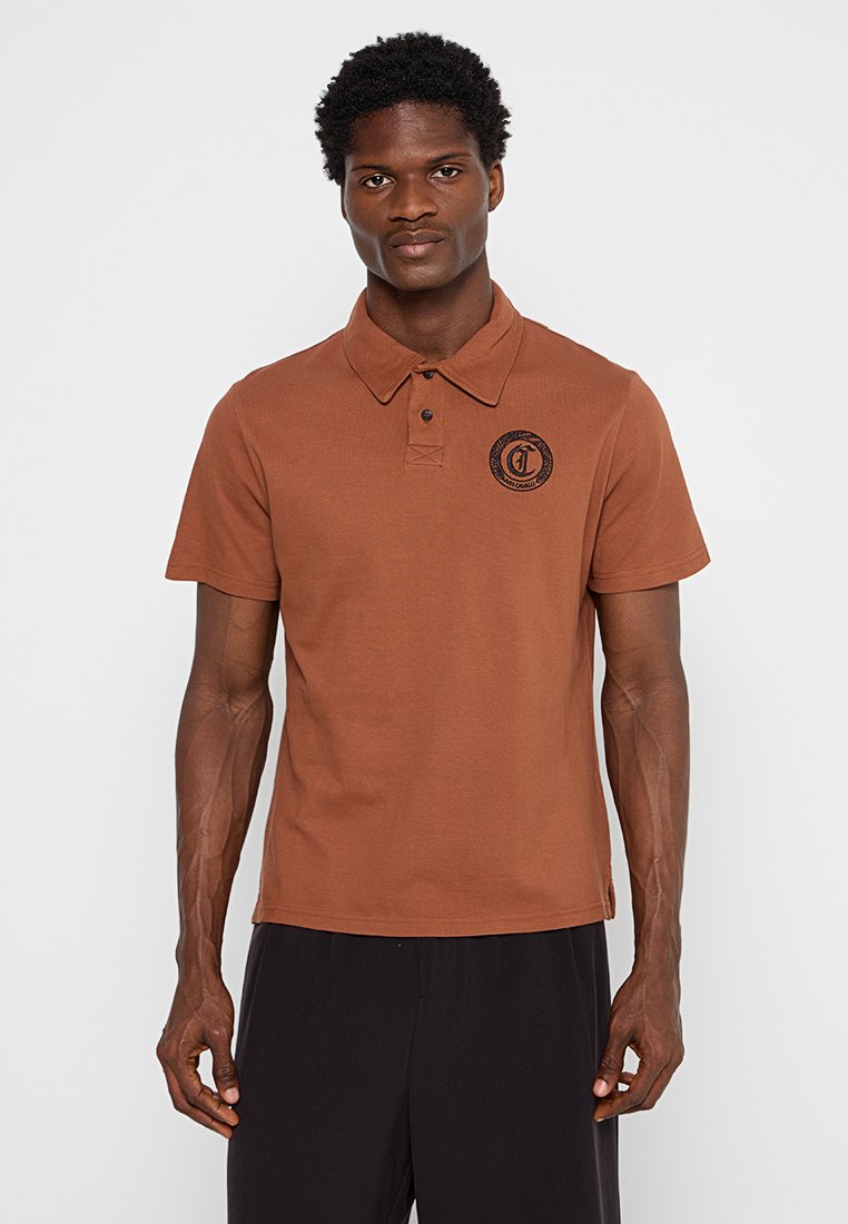 Braunes Poloshirt aus Baumwolle mit Kragen, kurzen Ärmeln und einem runden Logo auf der linken Brust. Glatte Textur und normale Passform.