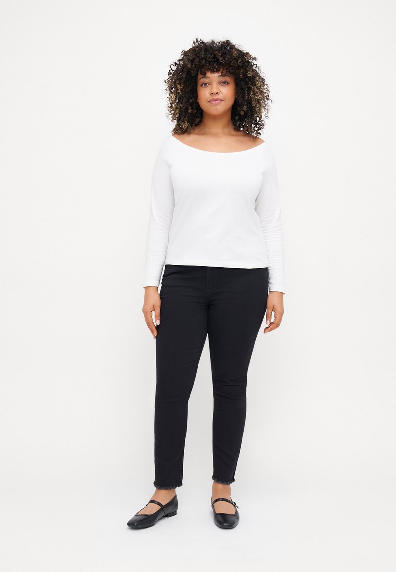 Weißes langärmeliges Off-Shoulder-Oberteil kombiniert mit schwarzen Skinny-Jeans mit ausgefransten Säumen und schwarzen Ballerinas mit Riemen über dem Spann.