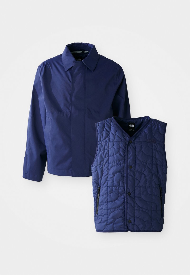 Marineblaue Herrenjacke mit Kragen und langen Ärmeln, neben einer gesteppte marineblauen ärmellosen Weste mit Druckknöpfen vorne und Reißverschlusstaschen.