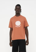 Mann trägt ein rostfarbenes T-Shirt mit einem weißen Kreis und dem Schriftzug "PS PAUL SMITH", kombiniert mit schwarzen Shorts, steht vor einem weißen Hintergrund.