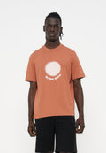 Mann trägt ein rostfarbenes T-Shirt mit einem weißen Kreis und dem Schriftzug "PS PAUL SMITH", kombiniert mit schwarzen Shorts, steht vor einem weißen Hintergrund.