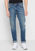 Frontansicht einer Person in hochgerollten blauen Jeans, einem schwarzen Shirt und weißen Sneakers, die vor einem einfarbigen hellen Hintergrund steht.