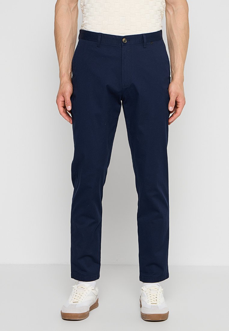Mann trägt dunkelblaue gerade geschnittene Hose und weiße Sneakers, steht vor einem schlichten hellen Hintergrund.
