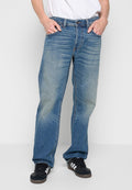 Mann in ausgewaschenen blauen gerade geschnittenen Jeans mit Händen in den Taschen und schwarzen Sneakers mit weißen Streifen, der vor einem weißen Hintergrund steht.