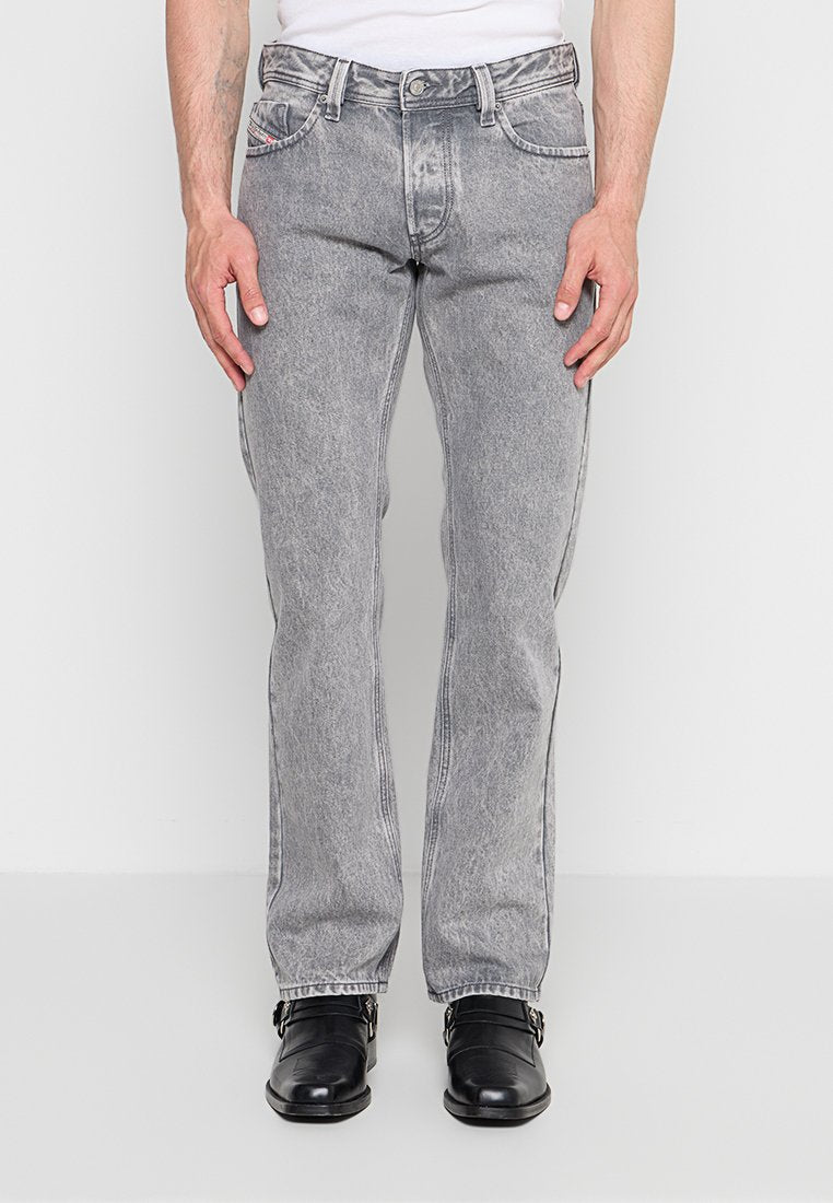 Mann trägt hellgraue, gerade Jeans mit weißem Hemd und schwarzen Lederschuhen mit Schnallen, steht vor einem einfarbigen Hintergrund.