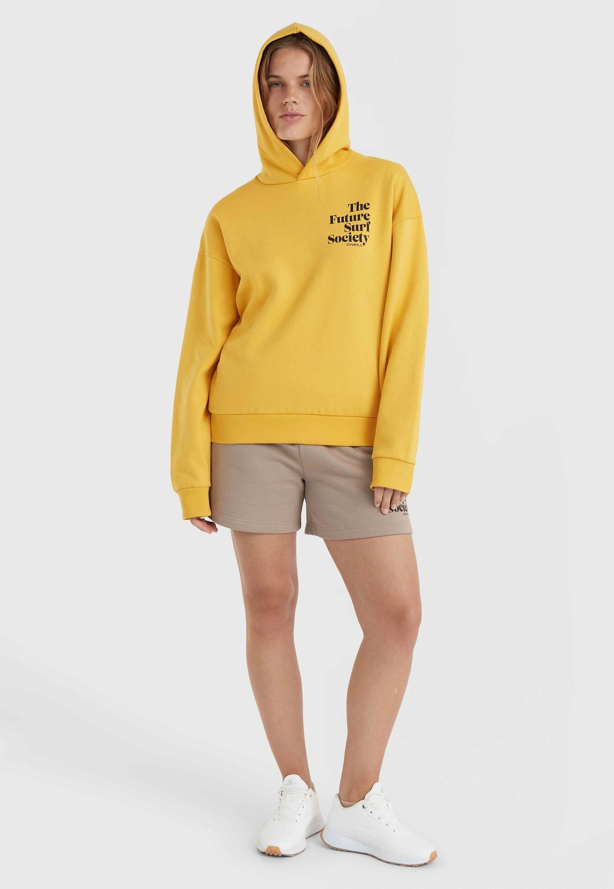 Gelbe Kapuzenjacke mit schwarzem Schriftzug, lockere Passform, Kapuze mit Kordelzug, kombiniert mit beigen Shorts und weißen Turnschuhen. Glatter Stoff, lässiger Stil.