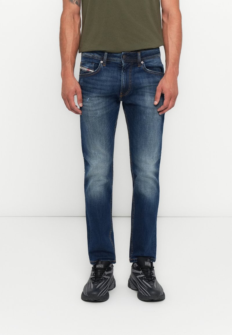 Mann in verblassten blauen Slim-Fit-Jeans, schwarzen Sneakers und einem olivgrünen Shirt, der vor einem schlichten weißen Hintergrund steht.