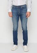 Mann in eng anliegenden blauen Jeans, weißen Sneakers und einem weißen Hemd mit Knöpfen, der vor einem schlichten Hintergrund steht.