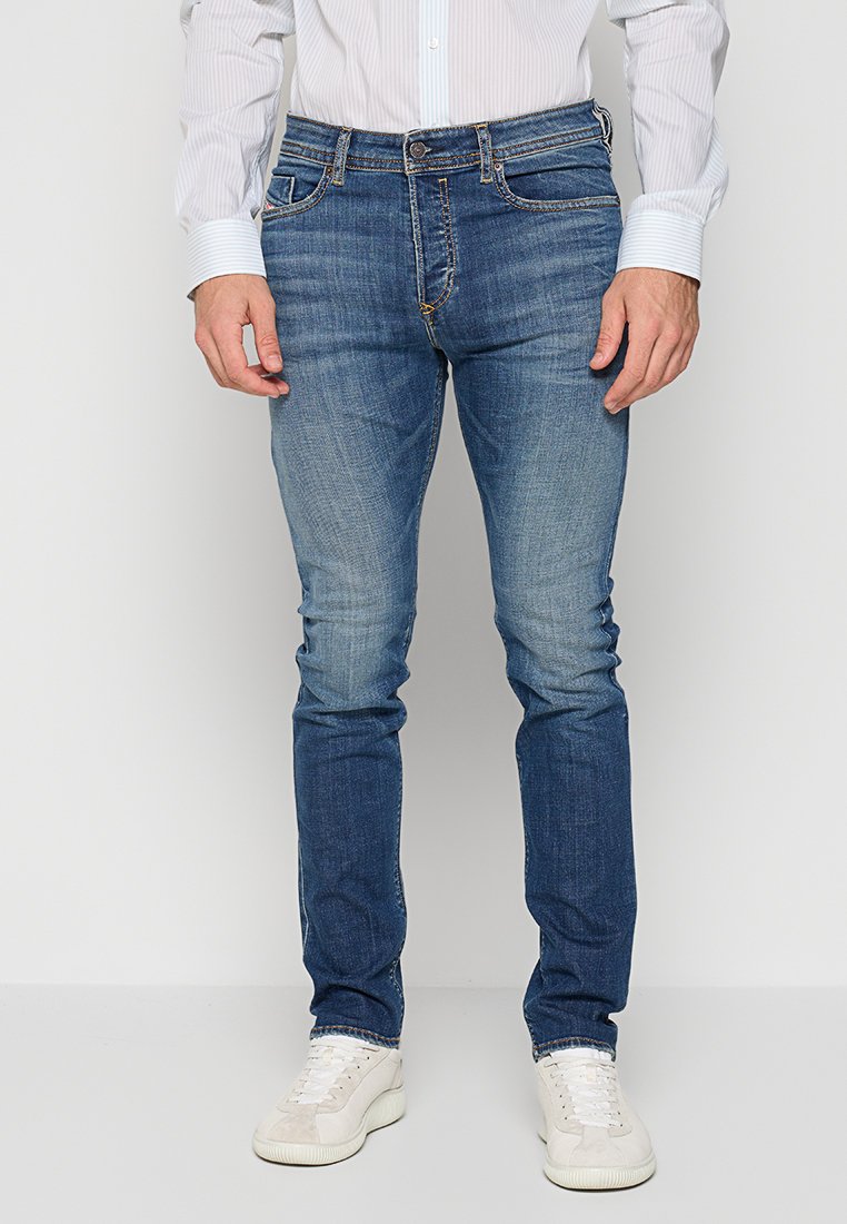 Mann in eng anliegenden blauen Jeans, weißen Sneakers und einem weißen Hemd mit Knöpfen, der vor einem schlichten Hintergrund steht.