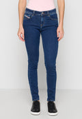 Frau trägt dunkelblaue Slim-Fit-Jeans, schwarze Sneakers und ein hellrosa ärmelloses Top, steht vor einem schlichten hellen Hintergrund.