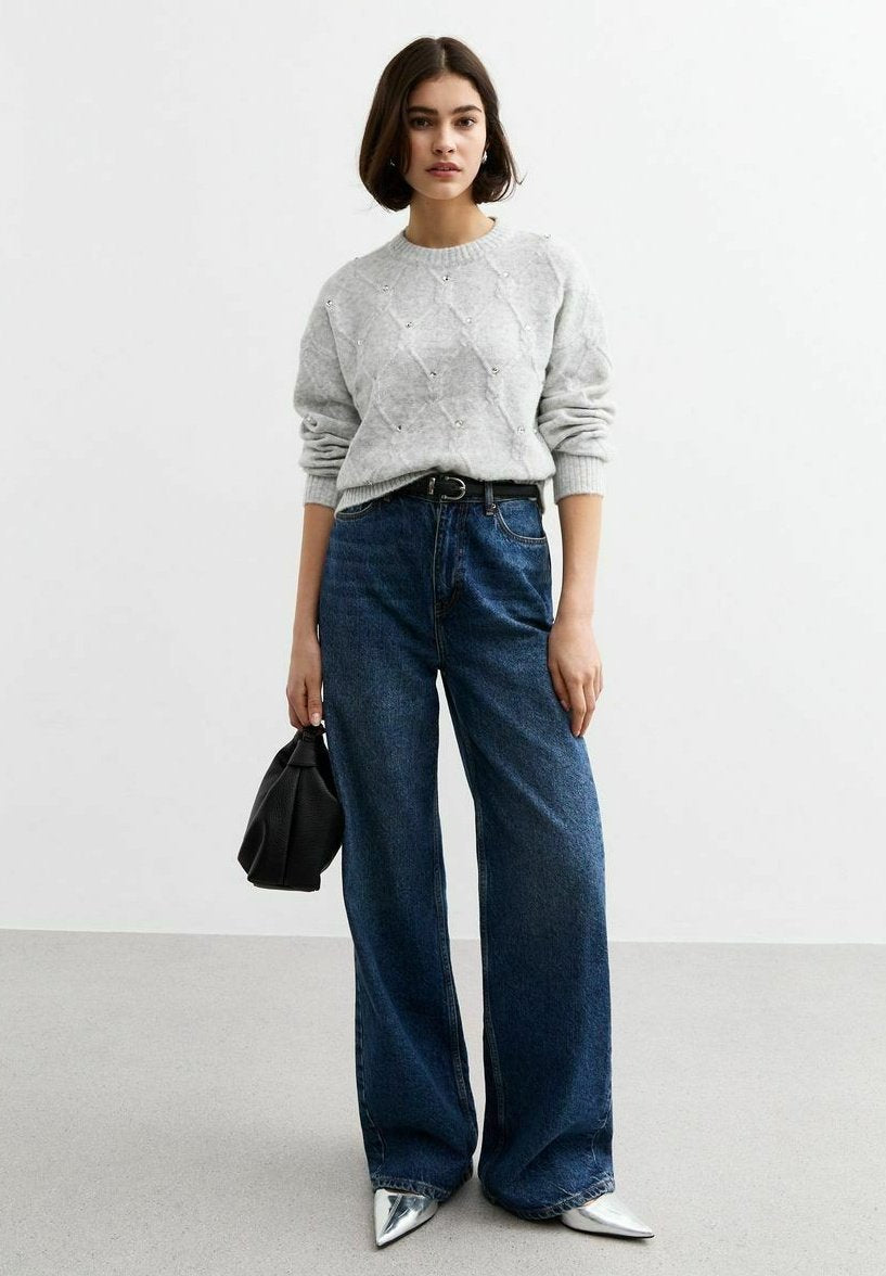 Grauer Strickpullover mit Rautenmuster und Verzierungen, kombiniert mit dunkelblauen weit geschnittenen Jeans. Mit einer schwarzen Handtasche und silbernen Absätzen accessoriert.