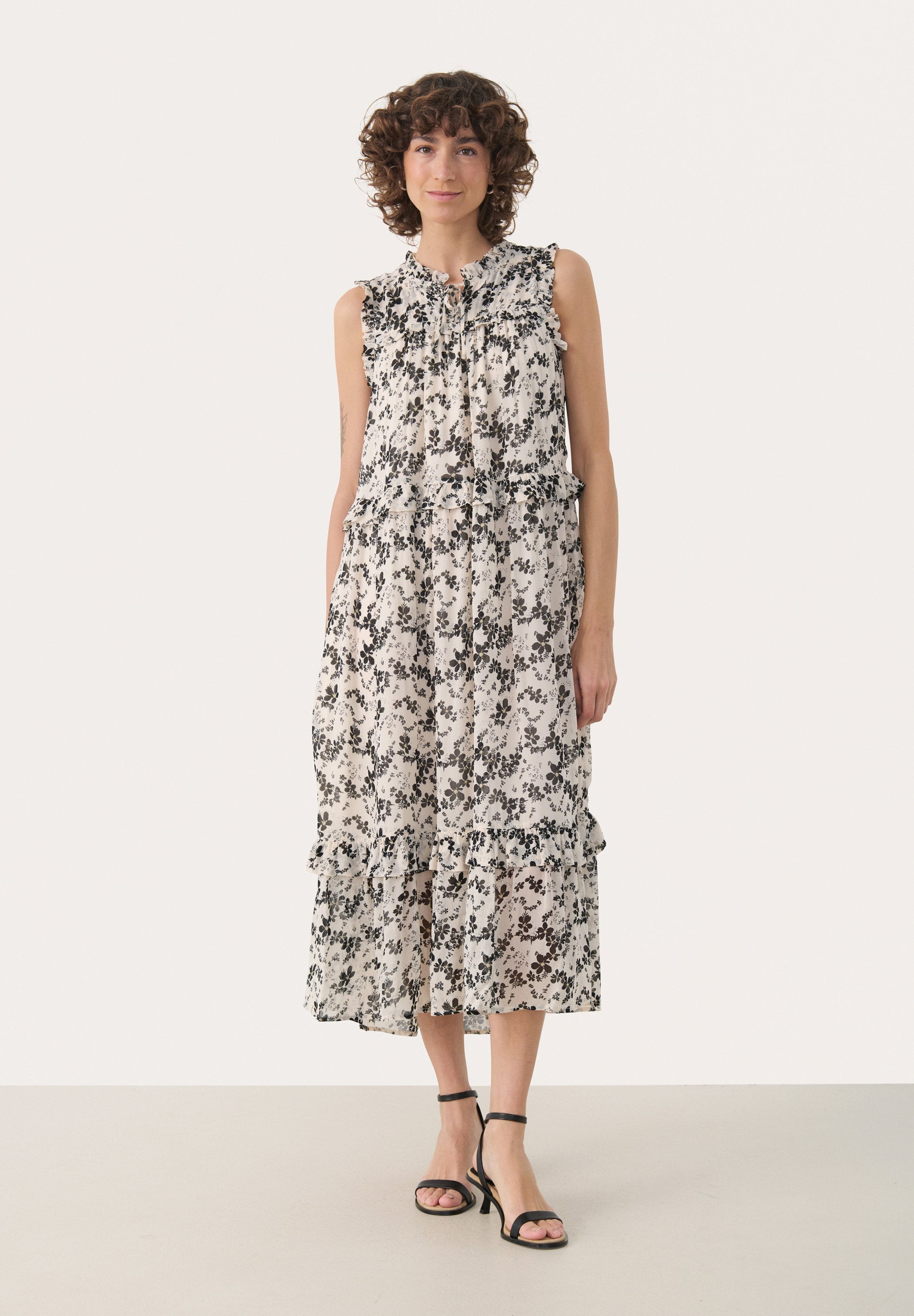 Ärmelloses Blumenkleid in Creme mit schwarzen Mustern, mit Rüschenakzenten und einem geschichteten Design. Kombiniert mit schwarzen Riemchensandalen.