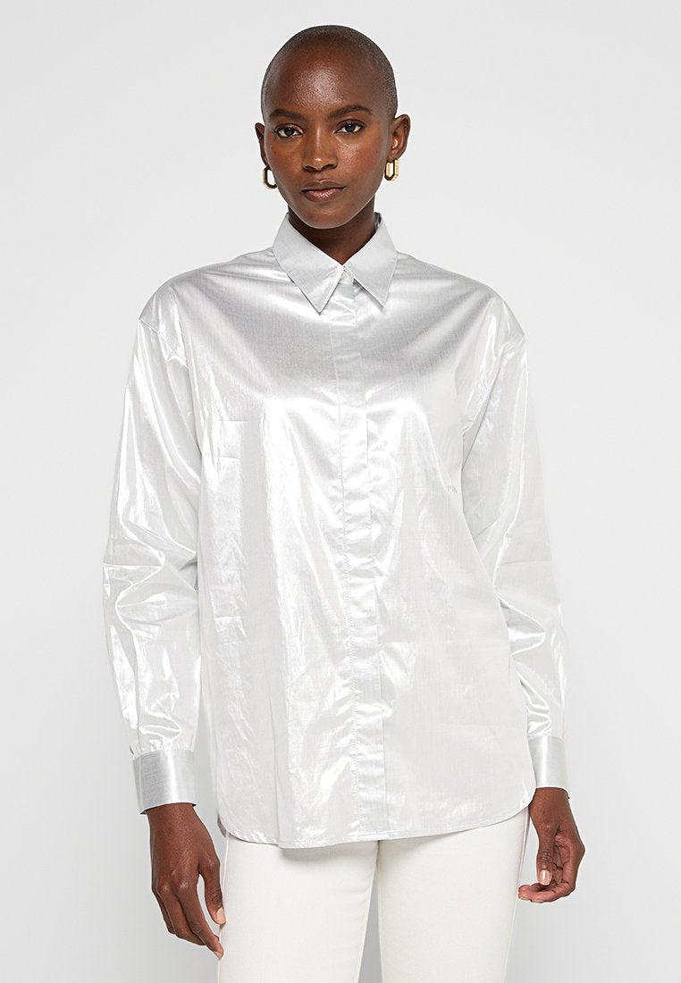Silberne, metallische Hemd mit Knopfverschluss, Kragen, langen Ärmeln und geradem Schnitt. Glatte Textur mit dezentem Glanz und einer Detailtasche auf der Vorderseite.