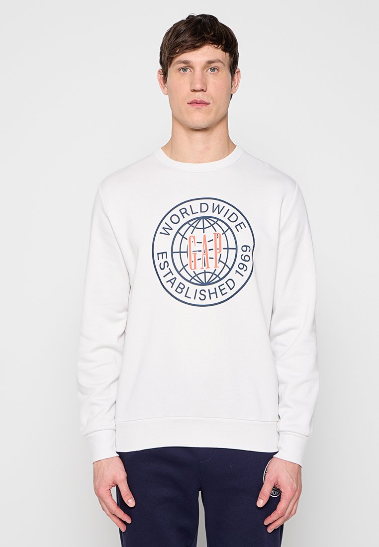 Junger Mann trägt ein weißes Sweatshirt mit blauem und orangefarbenem "GAP Worldwide Established 1969"-Logo und steht vor einem einfarbigen Hintergrund.