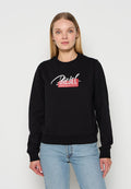 Frau mit glattem blondem Haar, die einen schwarzen Diesel-Sweatshirt und hellblaue Jeans trägt, vor einem weißen Hintergrund stehend.