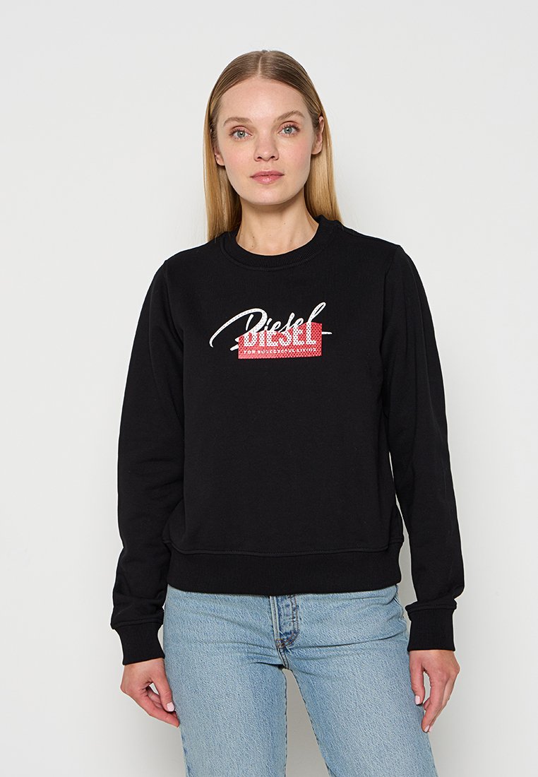Frau mit glattem blondem Haar, die einen schwarzen Diesel-Sweatshirt und hellblaue Jeans trägt, vor einem weißen Hintergrund stehend.