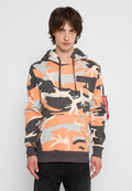 Junger Mann in einem orangefarbenen, grauen und beigen Camouflage-Hoodie mit Fronttasche und schwarzen Kordeln, der vor einem schlichten Hintergrund steht.