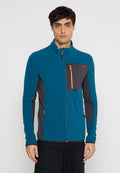 Mann in einer türkisfarbenen und dunkelgrauen Zip-Jacke mit orangefarbenen Reißverschlussakzenten, der vor einem schlichten hellen Hintergrund steht.