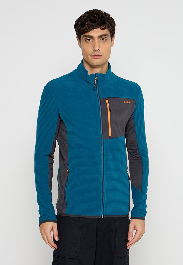 Mann in einer türkisfarbenen und dunkelgrauen Zip-Jacke mit orangefarbenen Reißverschlussakzenten, der vor einem schlichten hellen Hintergrund steht.