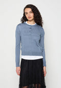 Frau mit lockigem dunklem Haar, die eine blaue, geknöpfte Strickjacke über einem weißen Oberteil und einem schwarzen Spitzenrock trägt, steht vor einem schlichten Hintergrund.