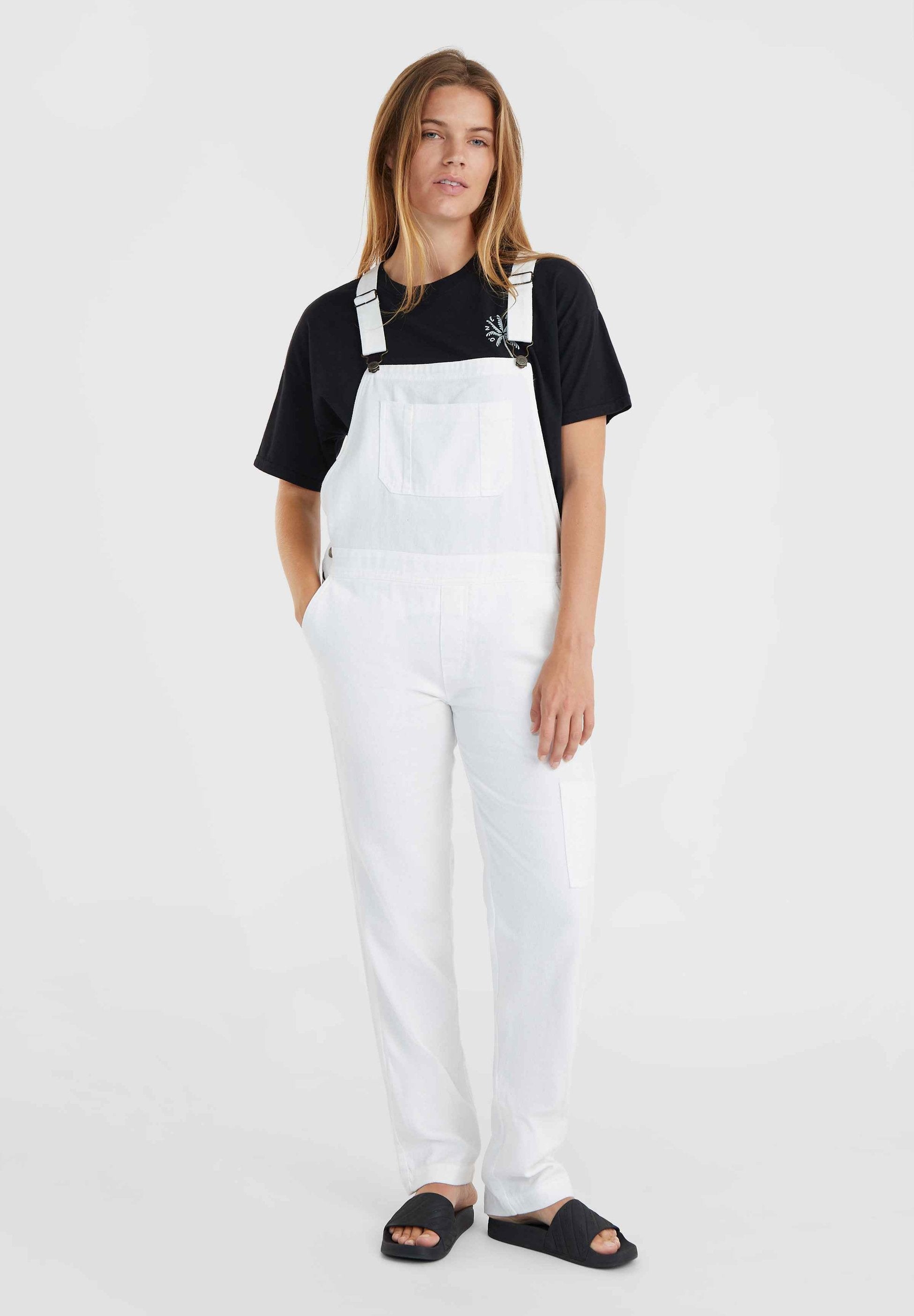 Weiße Denim-Overalls mit verstellbaren Trägern und einer Fronttasche, kombiniert mit einem schwarzen T-Shirt und schwarzen Badesandalen, in einem lässigen Schnitt.