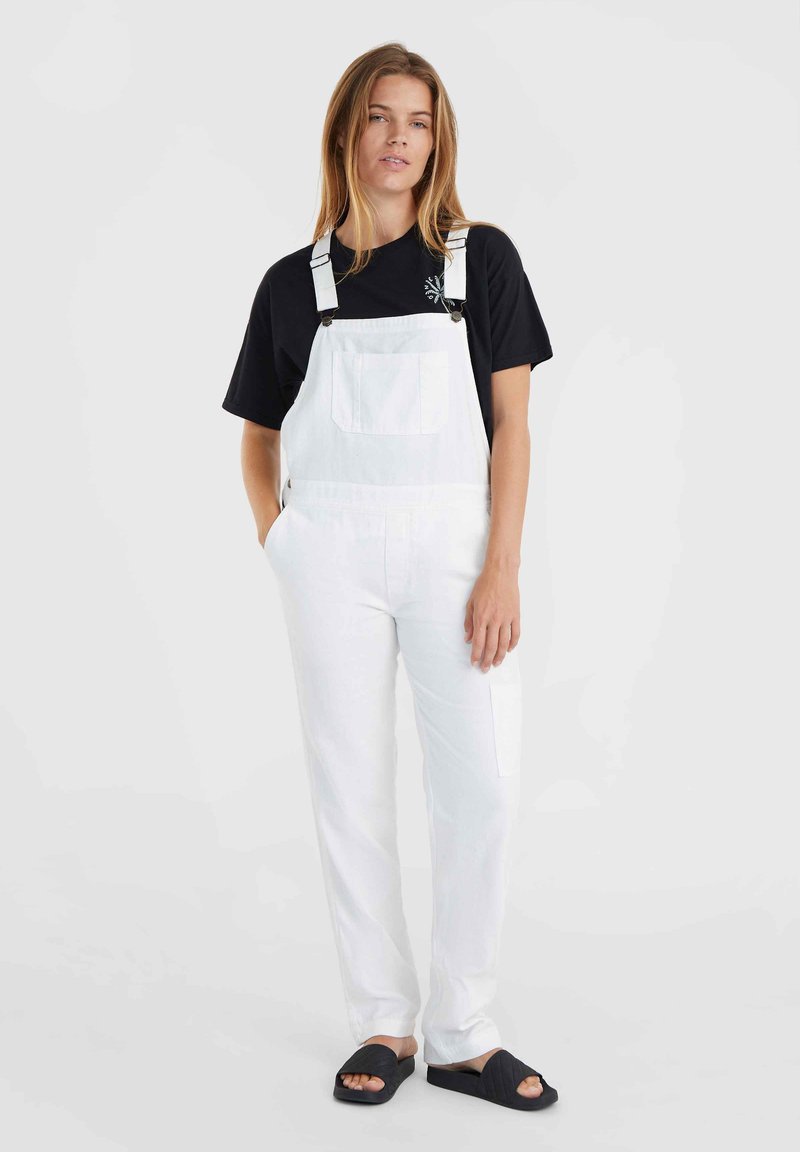 Weiße Denim-Overalls mit verstellbaren Trägern und einer Fronttasche, kombiniert mit einem schwarzen T-Shirt und schwarzen Badesandalen, in einem lässigen Schnitt.