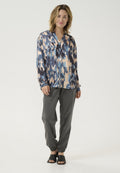 Seidenbluse mit Muster in Blau, Beige und Marineblau, mit V-Ausschnitt und Knopfleiste, kombiniert mit grauen Hose mit elastischem Bund und schwarzen Mules mit offenen Zehen.