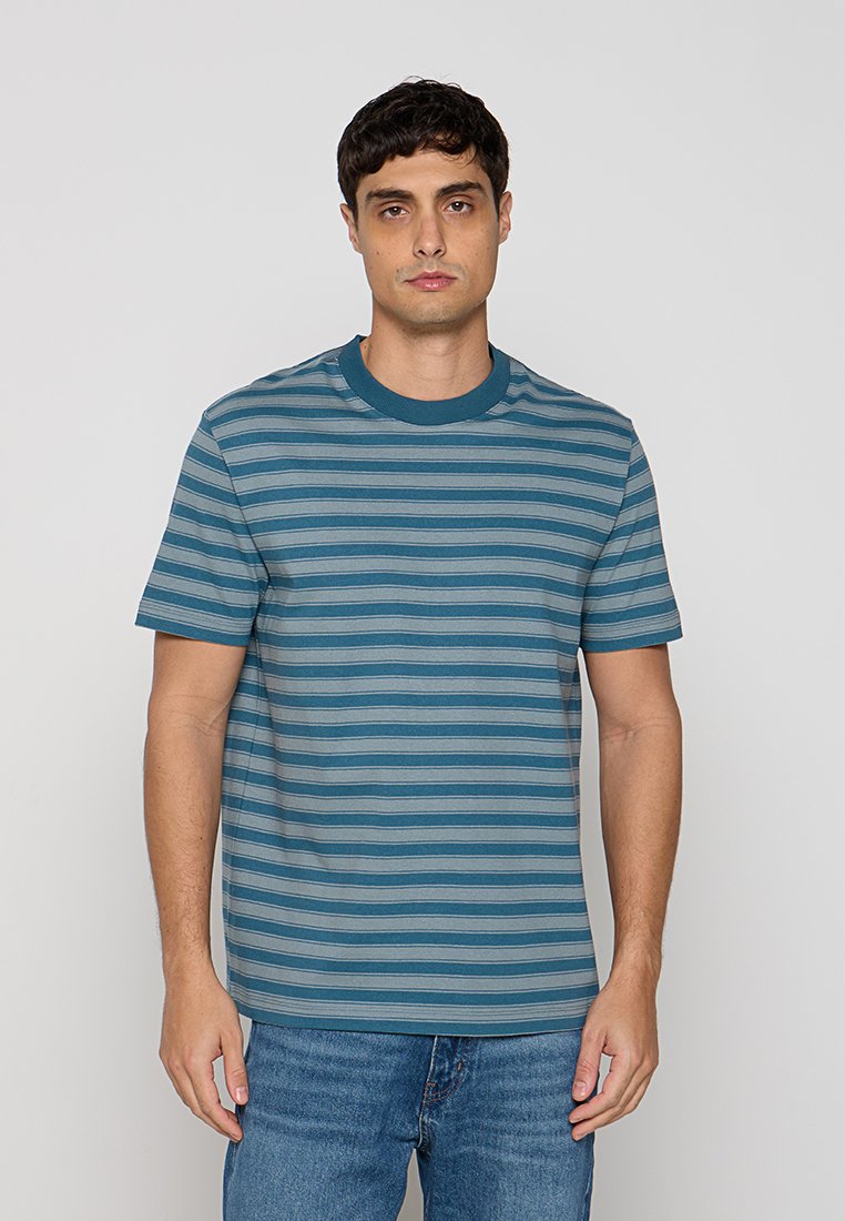 Junger Mann, der ein türkis-blau gestreiftes kurzärmeliges T-Shirt und blaue Jeans trägt, steht vor einem schlichten hellgrauen Hintergrund.