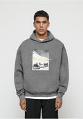 Junger Mann trägt einen grauen Hoodie mit Vintage-Auto-Grafik und dem Schriftzug „Peaks and Elegance“, steht vor einfachem, hellem Hintergrund.