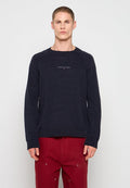 Mann trägt einen dunkelblauen, gesprenkelten Pullover mit "Tommy Jeans"-Logo und rote Hose, steht vor einem einfachen hellen Hintergrund.