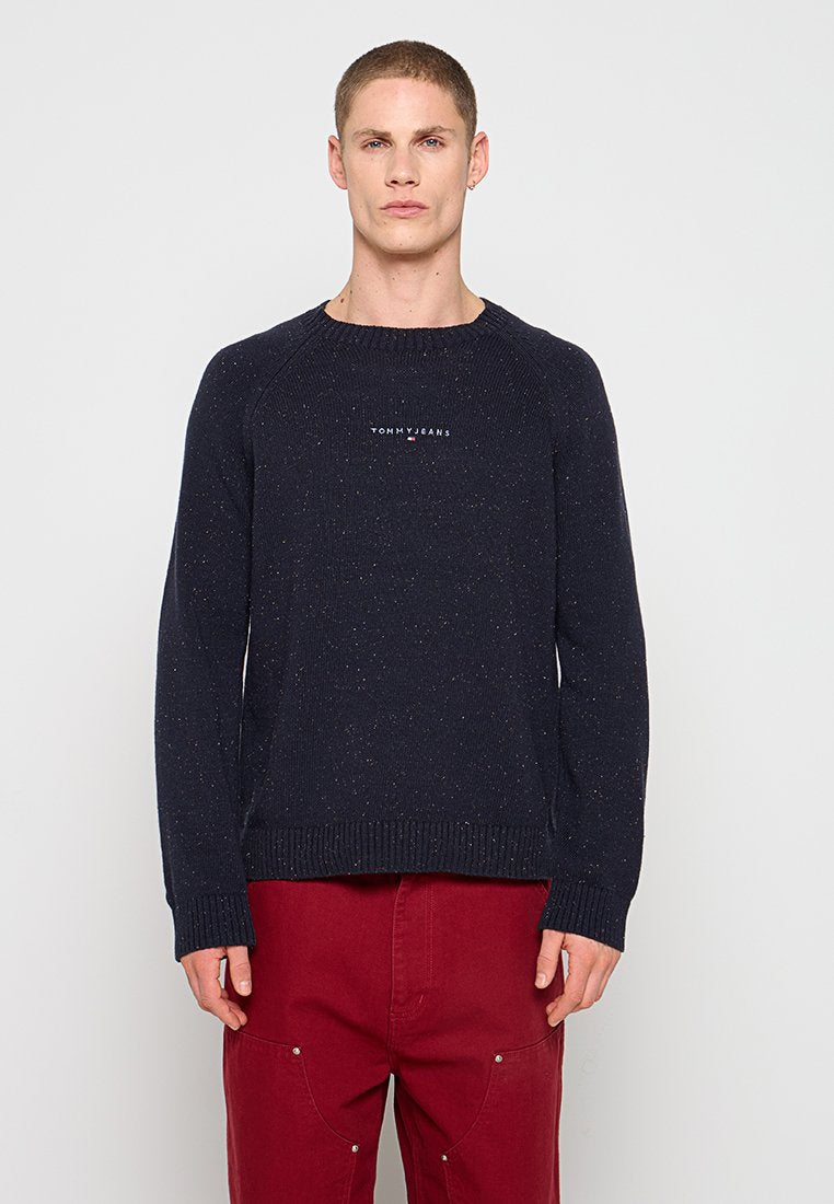 Mann trägt einen dunkelblauen, gesprenkelten Pullover mit "Tommy Jeans"-Logo und rote Hose, steht vor einem einfachen hellen Hintergrund.
