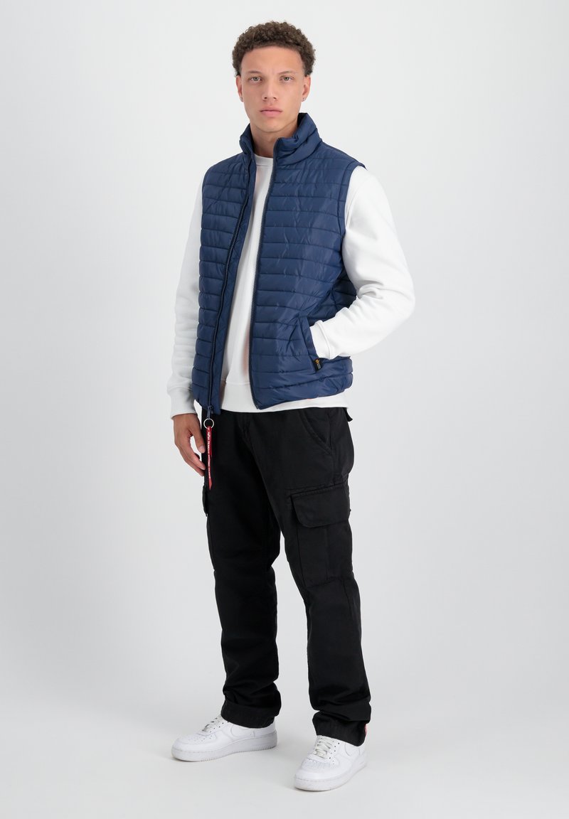 Marineblauer Steppweste mit Stehkragen, kombiniert mit einem weißen Sweatshirt, schwarzen Cargo-Hosen und weißen Sneakern; verfügt über seitliche Taschen und einen Reißverschluss.
