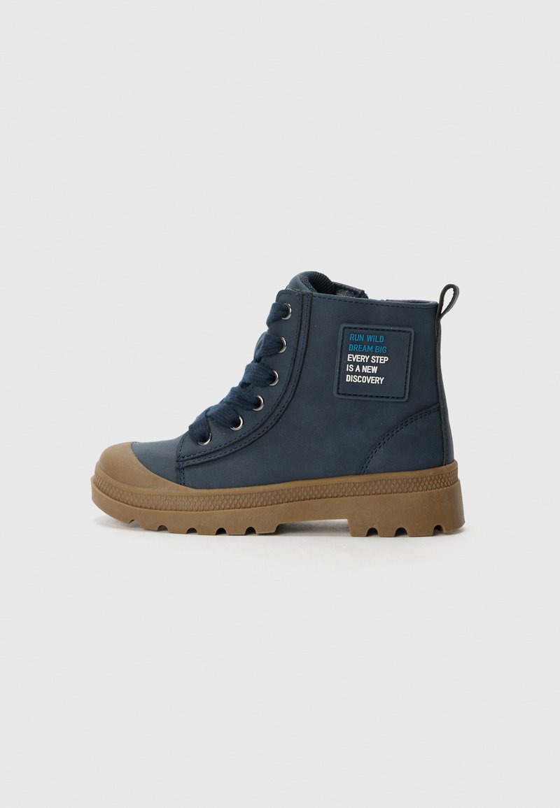 Marineblaue Knöchelstiefel aus synthetischem Material, mit brauner Gummisohle, runder Zehenform, sechs Metallösen und einem Patch mit Text.