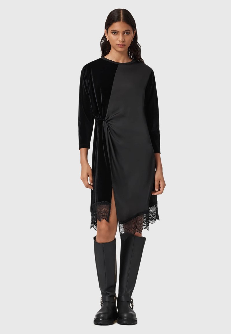 Schwarzes Samtkleid mit einem Knoten-Detail in der Taille und einer Spitzenkante am unteren Rand, mit langen Ärmeln und einem seitlichen Schlitz. Tragen mit hohen schwarzen Stiefeln.