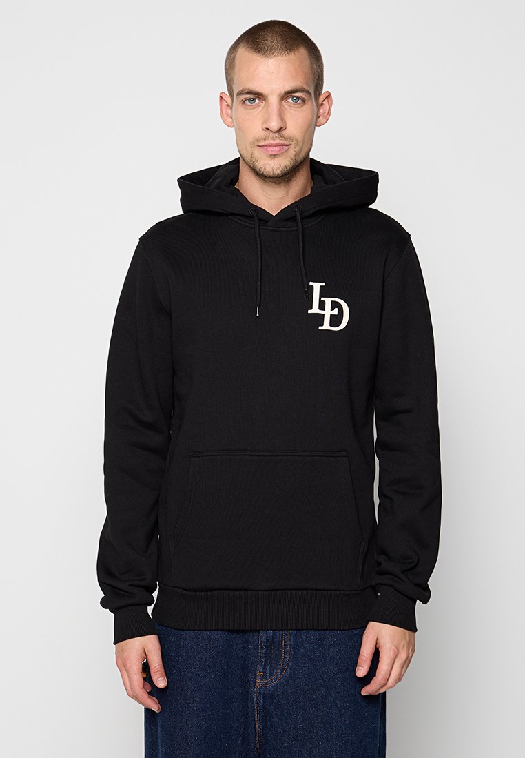 Mann in einem schwarzen Hoodie mit einer Vordertasche und "LD"-Logo auf der Brust, kombiniert mit blauen Jeans, steht vor einem einfarbigen Hintergrund.