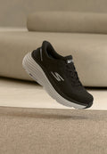 Schwarzer Skechers-Sneaker mit weißer, dicker "Max Cushion"-Sohle, der über einem beigen Teppich und einer Sofa-Kulisse schwebt.
