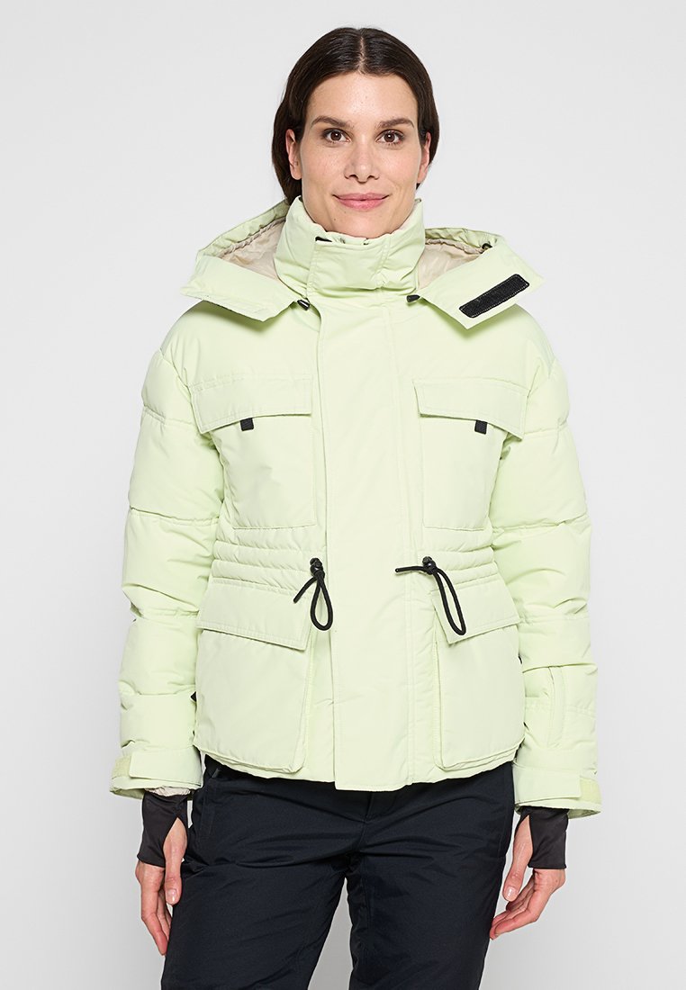 Hellgrüne Pufferjacke mit hohem Kragen, zwei Fronttaschen, Kordelzug in der Taille und beiger Innenauskleidung. Verstellbare Kapuze mit Hardware-Akzenten.