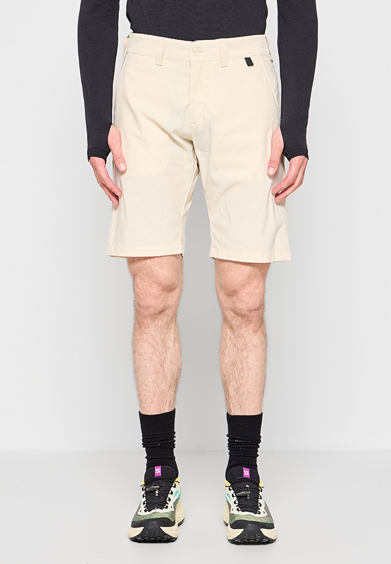 Mann trägt beige knielange Shorts, schwarzen Langarmoberteil, schwarze Socken und mehrfarbige Laufschuhe, steht vor einem schlichten Hintergrund.