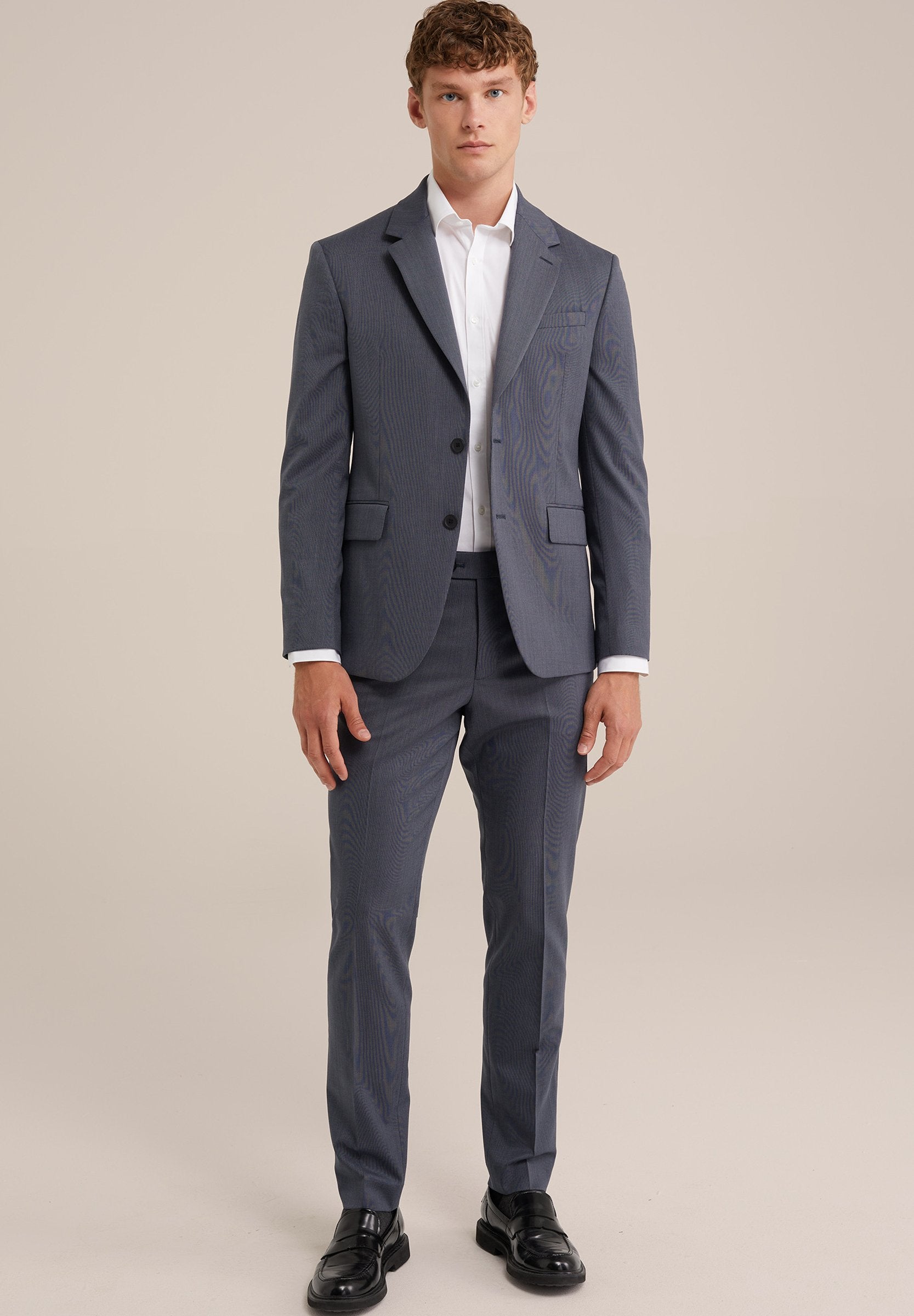 Grau gestreifter Anzug mit maßgeschneiderter Jacke und schmal geschnittenen Hosen. Darunter ein weißes Hemd. Schwarze Schuhe runden den formellen Look ab.
