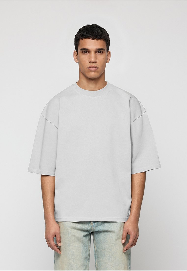 Mann steht vor einfarbigem Hintergrund, trägt ein übergroßes hellgraues kurzärmeliges T-Shirt und hellblaue Jeans, schaut mit neutralem Ausdruck nach vorne.
