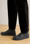 Schwarze Leder-Stiefeletten mit glatter Oberfläche, Schnürdesign und strukturiertem Gummisohle, kombiniert mit schwarzen Hosen auf einem Holzfußboden.