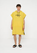 Junger Mann trägt einen gelben Kapuzenponcho mit schwarzen Kordeln und Logo, schwarze Slide-Sandalen, stehend vor weißem Hintergrund.