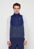 Junger Mann trägt eine marineblaue ärmellose Nike-Weste über einem weißen Langarmshirt vor einem einfarbigen hellen Hintergrund.